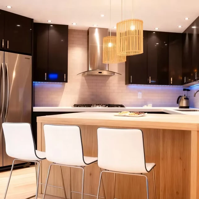 Modern custom kitchen design by Komandor GTA featuring a central light-wood kitchen island acting as a warm focal point, paired with three white contemporary bar stools with chrome frames. The perimeter cabinetry is finished in high-gloss black with sleek stainless steel hardware, contrasting sharply with the light grey subway tile backsplash and white countertops. Integrated cool-tone LED under-cabinet lighting illuminates the workspace, highlighting the texture of the tiles. Three cylindrical wood-slat pendant lights hang above the island, echoing the natural wood tones of the flooring. Stainless steel appliances, including a chimney-style range hood and gas cooktop, complete the high-end architectural aesthetic.