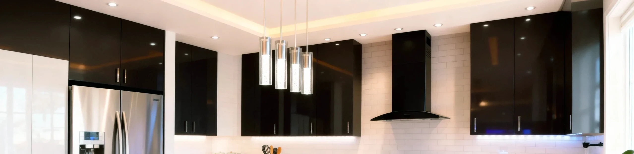 Modern custom kitchen design by Komandor GTA featuring floor-to-ceiling high-gloss black upper cabinetry with sleek stainless steel vertical bar handles. The installation utilizes integrated under-cabinet LED strip lighting to illuminate a classic white subway tile backsplash, enhancing the high-contrast minimalist aesthetic. Key appliances include a wall-mounted black chimney range hood and a stainless steel Samsung refrigerator seamlessly integrated into the joinery. Three contemporary cylindrical glass pendant lights hang from a drop ceiling with recessed pot lights, exemplifying a luxury European-inspired custom interior solution.